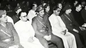 Group of men in military uniforms and traditional robes seated in an auditorium during a formal event (black-and-white photo).