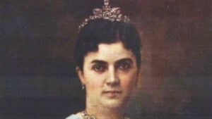 Portrait of a woman wearing a jeweled tiara and necklace, facing the camera in a formal setting.