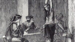 A woman bound to a vertical rack is suspended with wrists chained overhead as three men observe.