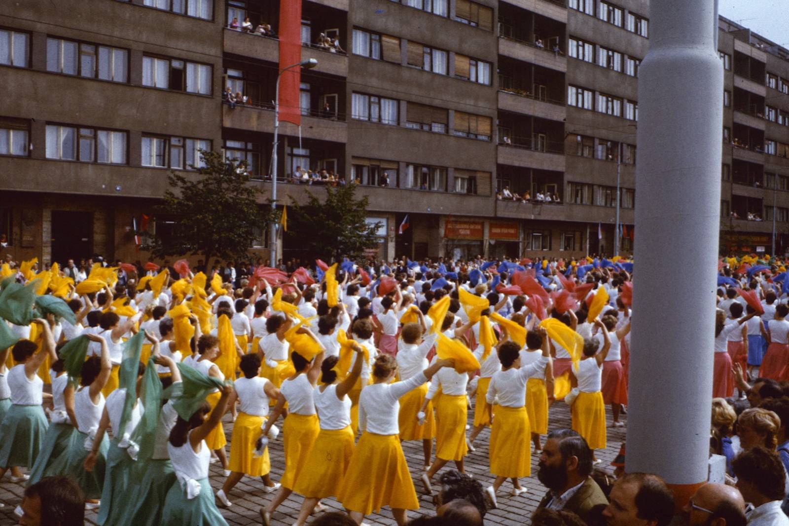 Crowd of performers in white T-shirts and yellow skirts waving colorful scarves during a city parade in front of apartment blocks.
