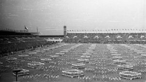 Large stadium field filled with thousands of people forming geometric drill formations during a parade or ceremony, with stands full of spectators in the background.