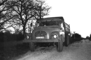 Front view of a vintage dump truck parked on a dirt road with leafless trees in the background.