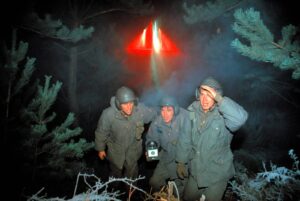 Three men in heavy coats and helmets crouch in a snowy forest at night beneath a red triangular light.