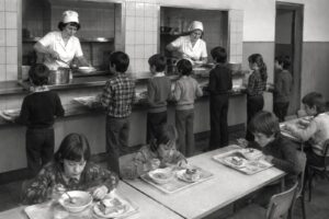 Children in a school cafeteria line up to be served by two cooks at a counter in trays and bowls